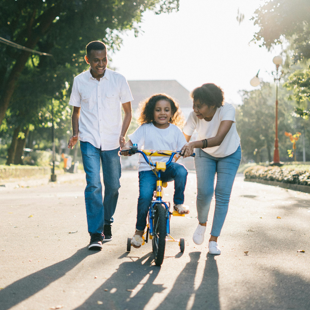 mother and father helping child learn how to ride a bike